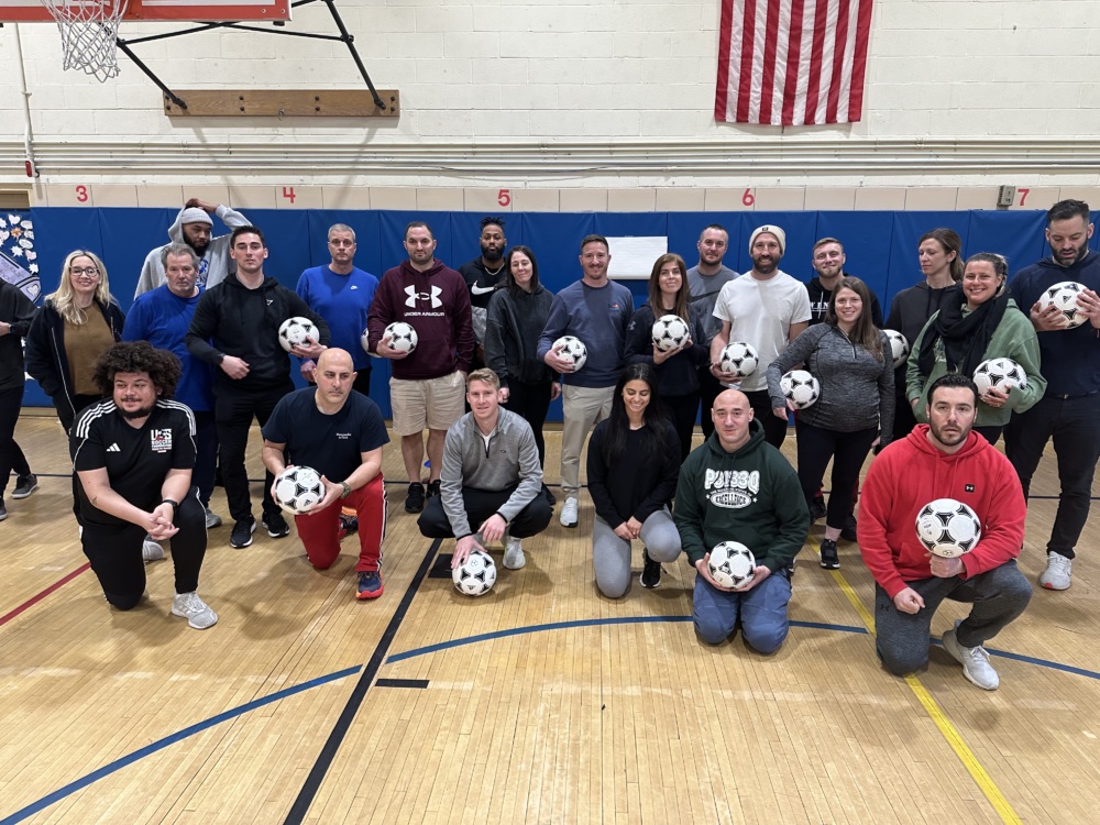 A group of PE teachers holding soccer balls pose for a photo in a school gym
